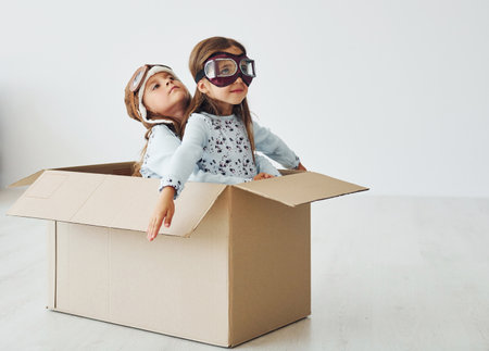 Sitting in the paper box. Two cute little girls indoors at home together. With retro pilot glasses and hatの写真素材