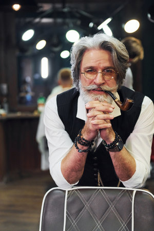 In retro clothes in barbershop. Stylish modern senior man with gray hair and beard is indoorsの写真素材