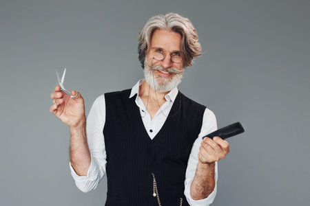 Professional barbershop worker. Against grey background. Stylish modern senior man with gray hair and beard is indoorsの写真素材