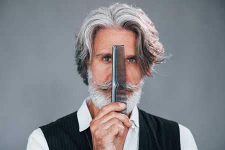 Professional barbershop worker. Against grey background. Stylish modern senior man with gray hair and beard is indoorsの写真素材
