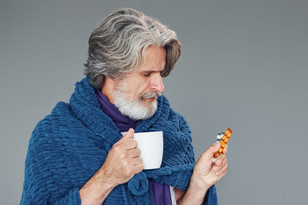 Getting sick. Against grey background. Stylish modern senior man with gray hair and beard is indoorsの写真素材