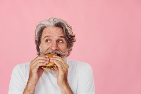 Eats delicious burger. Stylish modern senior man with gray hair and beard is indoorsの写真素材