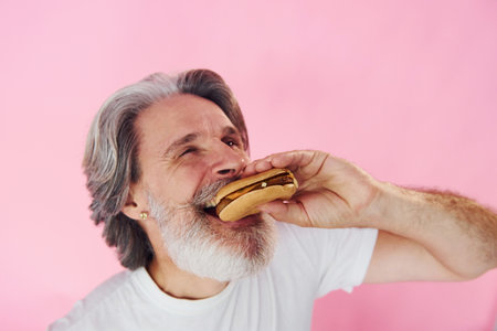 Eats delicious burger. Stylish modern senior man with gray hair and beard is indoorsの写真素材