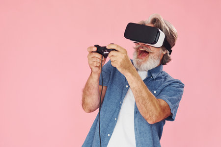 Playing game with virtual reality glasses and joystick. Stylish modern senior man with gray hair and beard is indoorsの写真素材