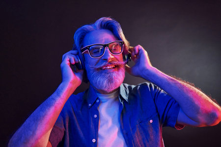Neon lighting. In headphones. Stylish modern senior man with gray hair and beard is indoors