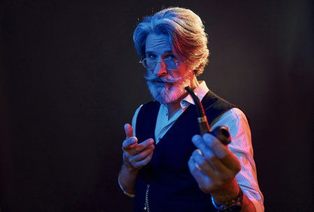Smoking and having fun. Neon lighting. Stylish modern senior man with gray hair and beard is indoorsの写真素材