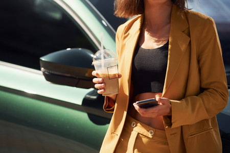 Holding drink and smartphone. Young fashionable woman in burgundy colored coat at daytime with her carの写真素材