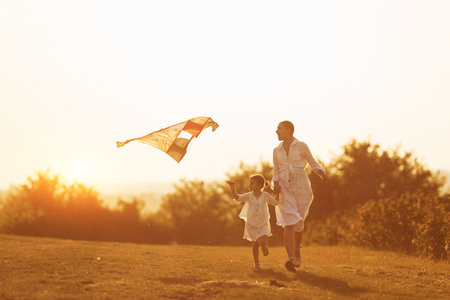 Daughter with her mother have fun together by running with kite on the field. Illuminated by beautiful sunlightの写真素材