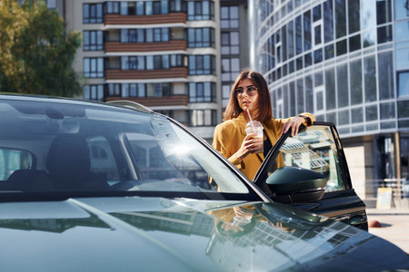 Against business building. Young fashionable woman in burgundy colored coat at daytime with her carの写真素材