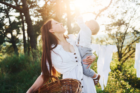 Clean clothes hanging on the rope to dry. Young mother with her little son is outdoors in the forest. Beautiful sunshineの写真素材