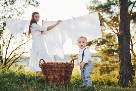 Clean clothes hanging on the rope to dry. Young mother with her little son is outdoors in the forest. Beautiful sunshineの写真素材