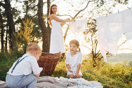 White clothes hanging on the rope to dry. Young mother with her little daughter and son is outdoors in the forest. Beautiful sunshineの写真素材
