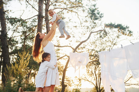 White clothes hanging on the rope to dry. Young mother with her little daughter and son is outdoors in the forest. Beautiful sunshineの写真素材
