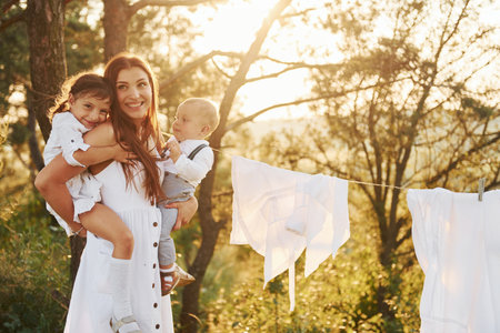 White clothes hanging on the rope to dry. Young mother with her little daughter and son is outdoors in the forest. Beautiful sunshineの写真素材