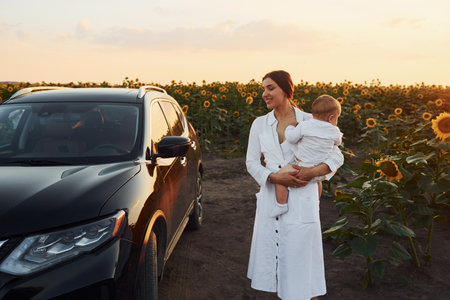 Near modern black car. Young mother with her little son is outdoors in the agricultural field. Beautiful sunshineの写真素材