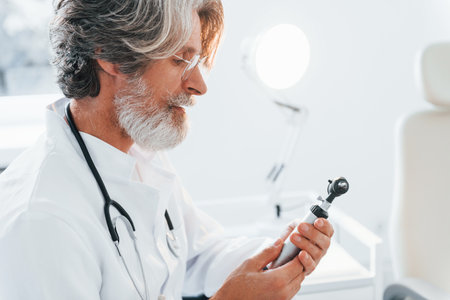 Portrait of senior male doctor with grey hair and beard in white coat is indoors in otorhinolaryngology clinicの写真素材