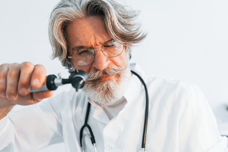 Portrait of senior male doctor with grey hair and beard in white coat is indoors in otorhinolaryngology clinicの写真素材