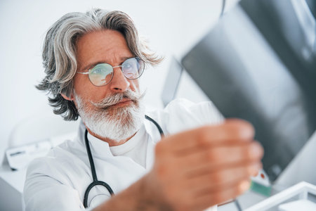 Looks at x-ray. Senior male doctor with grey hair and beard in white coat is indoors in clinic