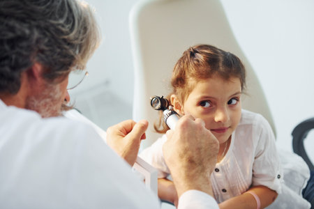 Little girl have a visit. Senior otorhinolaryngologist with grey hair and beard in white coat is indoors in clinicの写真素材