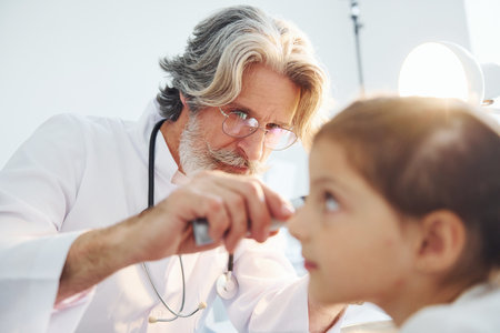 Little girl have a visit. Senior otorhinolaryngologist with grey hair and beard in white coat is indoors in clinicの写真素材