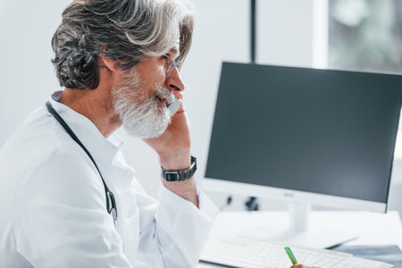 Works by computer table. Senior male doctor with grey hair and beard in white coat is indoors in clinicの写真素材