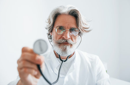 Holds stethoscope. Portrait of senior male doctor with grey hair and beard in white coat is indoors in clinicの写真素材