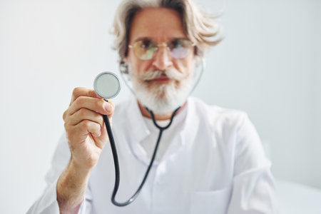 Holds stethoscope. Portrait of senior male doctor with grey hair and beard in white coat is indoors in clinicの写真素材