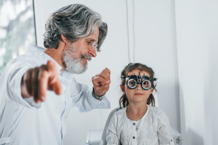 Senior male ophthalmologist with grey hair and beard in white coat is indoors in clinic testing sight of little girlの写真素材