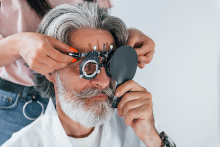 Getting tested by woman worker. Senior man with grey hair and beard is in ophthalmology clinicの写真素材