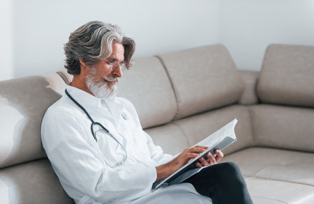 Senior male doctor with grey hair and beard in white coat is indoors in clinic sits on sofa with notepad in handsの写真素材