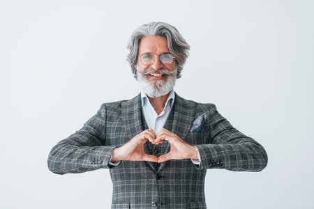 In elegant clothes. Standing against white wall. Senior stylish modern man with grey hair and beard indoorsの写真素材