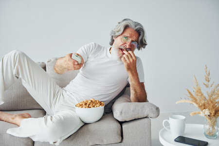 Watching TV show and eats popcorn. Senior stylish modern man with grey hair and beard indoorsの写真素材