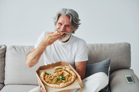 Eats delicious pizza while watching TV show. Senior stylish modern man with grey hair and beard indoorsの写真素材