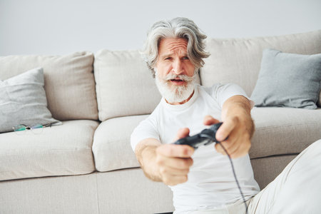 Plays video game by using controller. Senior stylish modern man with grey hair and beard indoorsの写真素材