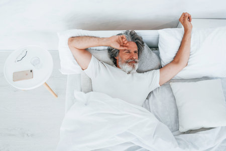 Senior stylish modern man with grey hair and beard indoors lying on the bed and waking upの写真素材