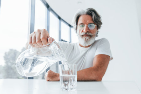 Pours water into glass. Senior stylish modern man with grey hair and beard indoorsの写真素材