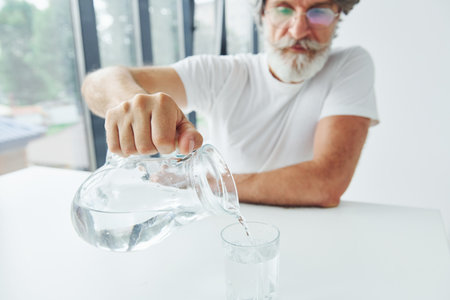 Pours water into glass. Senior stylish modern man with grey hair and beard indoorsの写真素材