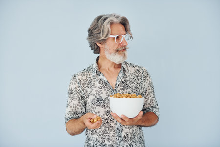 Standing with popcorn against wall. Senior stylish modern man with grey hair and beard indoorsの写真素材