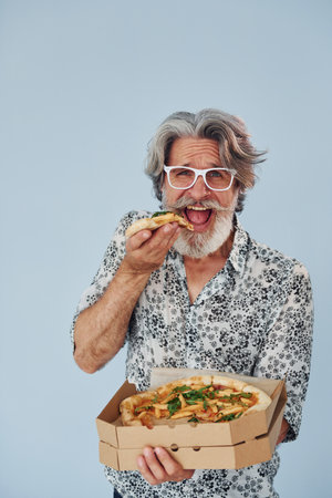 Eats pizza when standing against wall. Senior stylish modern man with grey hair and beard indoorsの写真素材