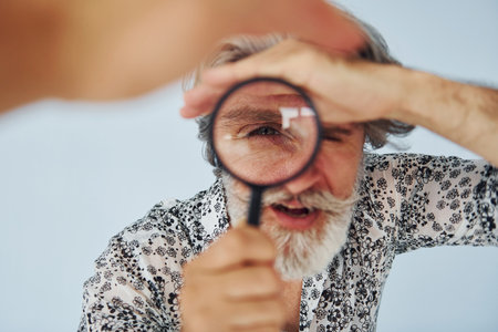 Having fun with magnifying glass. Senior stylish modern man with grey hair and beard indoorsの写真素材