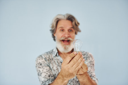 Holds light bulb in hands. Conception of ideas and inspiration. Senior stylish modern man with grey hair and beard indoorsの写真素材