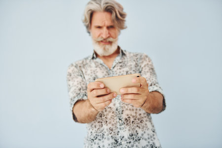 Playing game on phone. Senior stylish modern man with grey hair and beard indoorsの写真素材