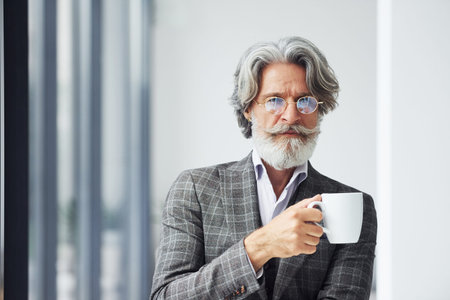 Takes a break and drinks coffee. Senior stylish modern man with grey hair and beard indoorsの写真素材