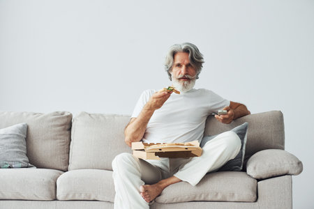 With delicious pizza. Watches TV show. Senior stylish modern man with grey hair and beard indoorsの写真素材