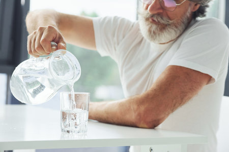Senior stylish modern man with grey hair and beard indoors pouring water into glassの写真素材