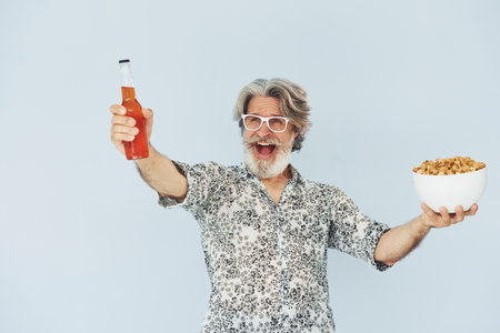 Senior stylish modern man with grey hair and beard indoors with bottle of drink and popcornの写真素材