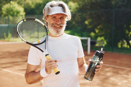 Taking a break and drinking water. Senior modern stylish man with racket outdoors on tennis court at daytimeの写真素材