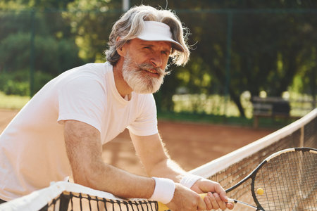Senior modern stylish man with racket outdoors on tennis court at daytimeの写真素材