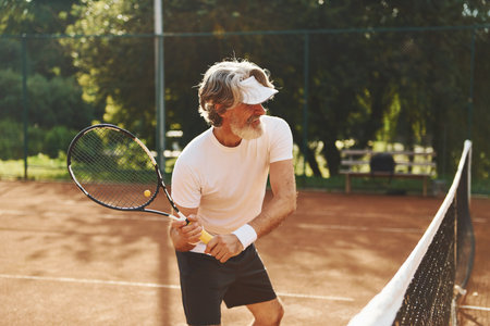 Playing game. Senior modern stylish man with racket outdoors on tennis court at daytimeの写真素材