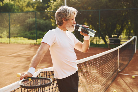 Taking a break and drinking water. Senior modern stylish man with racket outdoors on tennis court at daytimeの写真素材
